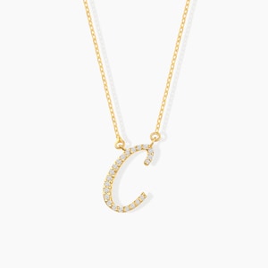 May include: A gold-colored necklace featuring the letter "C" encrusted with small, sparkling gemstones. The pendant hangs from a delicate chain. The necklace is displayed against a plain white background.