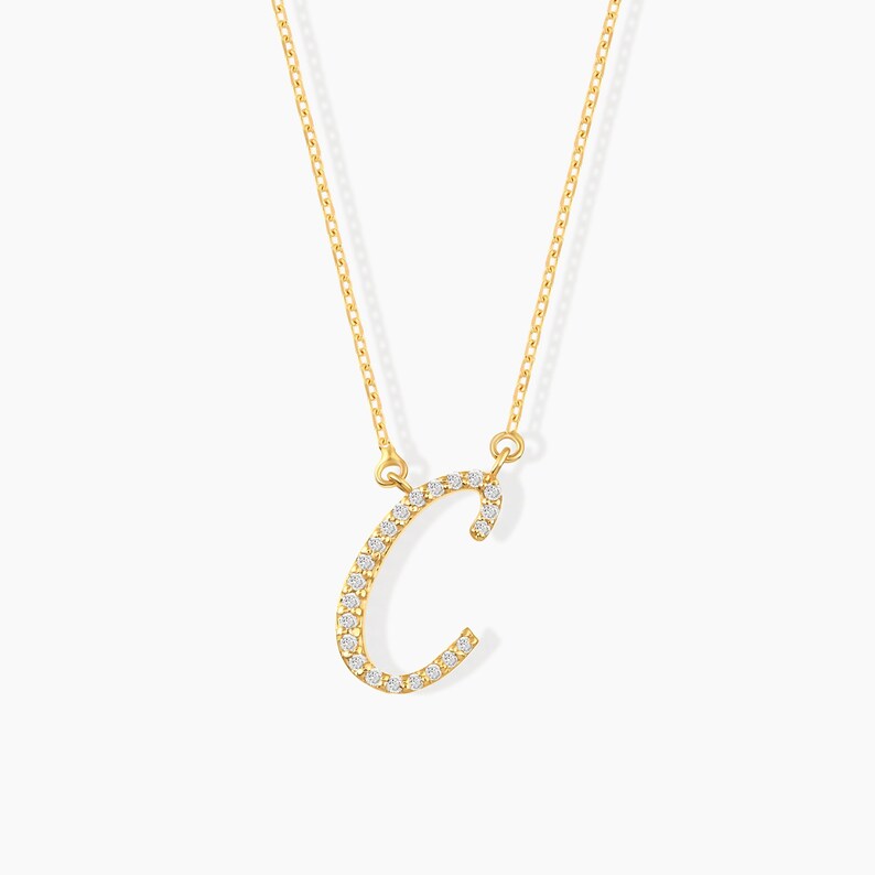 May include: A gold-colored necklace featuring the letter "C" encrusted with small, sparkling gemstones. The pendant hangs from a delicate chain. The necklace is displayed against a plain white background.