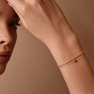 May include: Two gold bracelets on a wrist. One is a delicate chain, the other a rope design. A small red heart charm hangs from the chain bracelet. The background is a solid, neutral color.