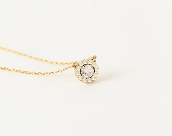 May include: A delicate gold chain necklace with a small round pendant featuring a single clear stone surrounded by a halo of smaller stones.