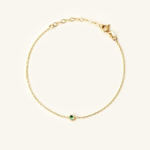 May include: A delicate gold chain bracelet featuring a single, small, round, bezel-set green gemstone centered on the chain. The bracelet includes a thin, polished gold link chain with a small lobster claw clasp and an adjustable extender section.