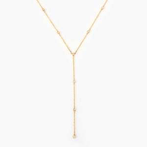 Pode incluir: Um colar de corrente de ouro com pequenas pedras transparentes. O colar é de estilo lariat, com uma corrente longa e fina que cai na frente do pescoço.