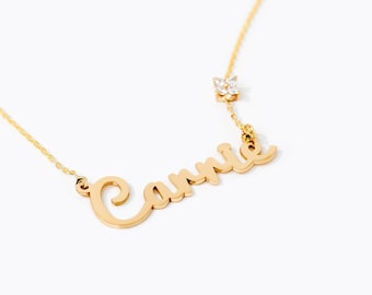 May include: A gold-toned necklace featuring a cursive nameplate pendant that reads "Carrie" in a flowing script font. A small, four-petaled flower-shaped charm set with clear, sparkling stones is attached to the chain just above the nameplate.