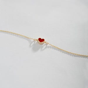May include: A delicate gold chain bracelet featuring a small, red heart charm. The heart is centered on the chain and has a smooth, polished surface. The bracelet is displayed on a white background.