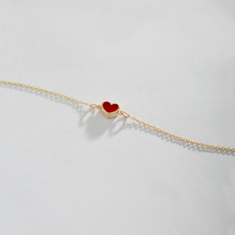 May include: A delicate gold chain bracelet featuring a small, red heart charm. The heart is centered on the chain and has a smooth, polished surface. The bracelet is displayed on a white background.