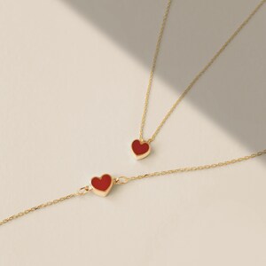 May include: A gold-colored necklace and bracelet set, each featuring a red heart-shaped pendant. The jewelry is displayed on a light-colored surface, with soft shadows adding depth. The heart pendants have a smooth, polished finish.