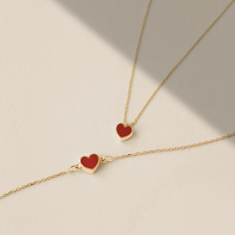 May include: A gold-colored necklace and bracelet set, each featuring a red heart-shaped pendant. The jewelry is displayed on a light-colored surface, with soft shadows adding depth. The heart pendants have a smooth, polished finish.