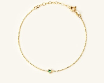 May include: A delicate gold chain bracelet featuring a single, small, round, bezel-set green gemstone centered on the chain. The bracelet includes a thin link chain, a circular spring ring clasp, and an adjustable length section with small circular links.