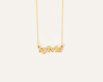May include: A gold-toned necklace featuring a delicate chain and a central pendant displaying the name Sylvie in a lowercase, sans-serif font. The metal has a smooth, reflective finish, and the pendant is securely attached to the chain on both sides.