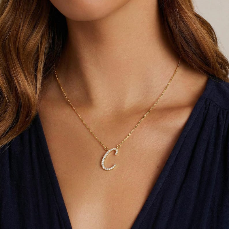 May include: A gold-colored necklace with a pendant in the shape of the letter "C". The pendant is encrusted with small, clear crystals. The necklace is displayed on a person wearing a dark blue top.