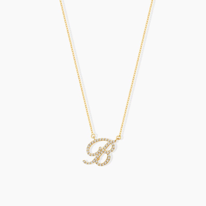 May include: A delicate gold necklace featuring the letter "B" encrusted with small, sparkling gemstones. The pendant hangs from a fine gold chain, creating a simple yet elegant piece of jewelry. The necklace is displayed against a plain white background.