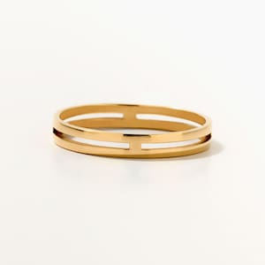 May include: A gold-toned ring featuring a smooth, polished finish and a distinctive open-work design. The band consists of two parallel horizontal bars connected by small vertical segments, creating a clean, geometric, and continuous circular structure.