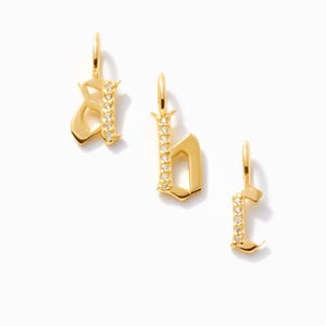 May include: Three gold-toned metal letter pendants in an old English font, each featuring a vertical row of small, clear, sparkling stones set along one side. Each pendant includes a small circular jump ring at the top for attachment to a chain.