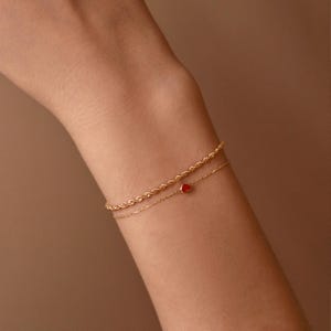 May include: Two delicate gold bracelets. The top bracelet has a rope chain design. The bottom bracelet features a small red heart charm. The bracelets are worn on a person's arm.