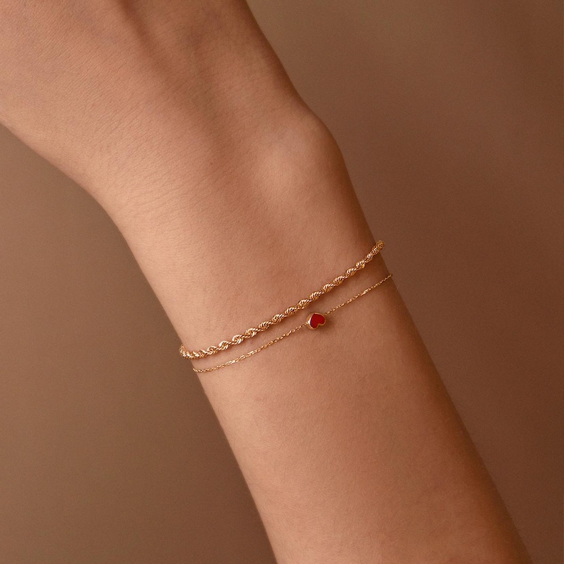 May include: Two delicate gold bracelets. The top bracelet has a rope chain design. The bottom bracelet features a small red heart charm. The bracelets are worn on a person's arm.