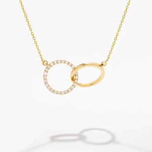 May include: A gold necklace featuring two interlocking circular pendants. One circle is encrusted with small, clear, round-cut stones, while the other is a plain, polished gold band. The piece hangs from a delicate gold chain with small links.