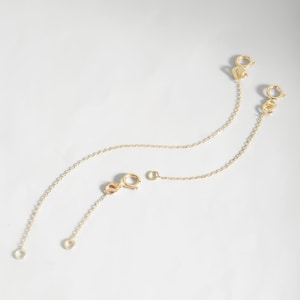 May include: Two gold-toned metal jewelry extender chains, each featuring a delicate cable link design. One end of each chain is finished with a circular spring ring clasp, while the other end terminates in a small, open jump ring connector.