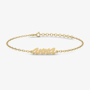 May include: A gold-toned chain bracelet featuring a central nameplate with the name Anna written in a cursive script font. The bracelet includes a delicate link chain and a small circular clasp mechanism at the end for secure fastening.