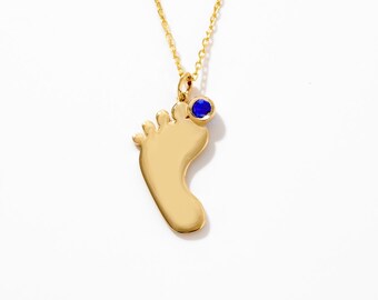 May include: A gold-toned necklace featuring a pendant shaped like a human foot. The pendant includes a small, round, deep blue gemstone set near the ankle area. The piece hangs from a delicate gold-toned chain with fine, uniform links.