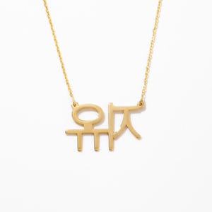May include: A gold-toned necklace featuring a pendant with two distinct Korean Hangul characters. The pendant is suspended from a delicate gold-toned chain. The design consists of clean, linear metal cutouts forming the characters, creating a polished and reflective surface finish throughout the entire piece of jewelry.