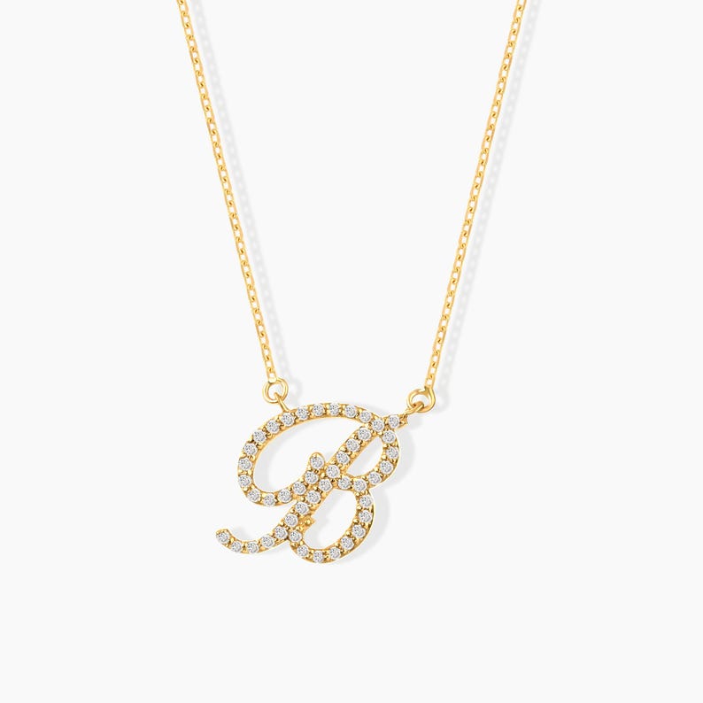 May include: A gold-colored necklace featuring the letter "B" encrusted with small, clear gemstones. The pendant is suspended from a delicate gold chain. The necklace is displayed against a white background.