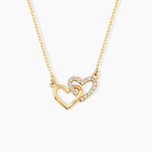 May include: A gold-toned necklace featuring two interlocking heart-shaped pendants. One heart is plain polished gold, while the other is encrusted with small, clear, sparkling stones. The pendant hangs from a delicate, thin gold-toned chain.
