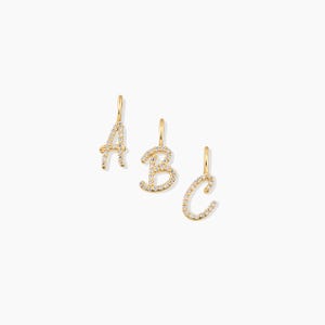 May include: Three gold-coloured initial charms spelling "A", "B", and "C". Each charm is encrusted with small, clear gemstones. The letters are in a cursive font, with a small loop at the top for hanging.