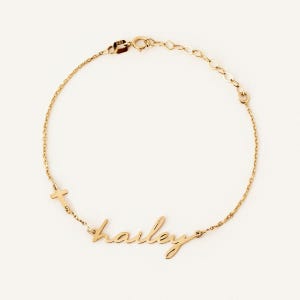 May include: A gold chain bracelet featuring a delicate script nameplate that reads Hailey, accompanied by a small cross charm. The bracelet includes a thin link chain, a secure clasp, and a small metal tag stamped with the number 585.