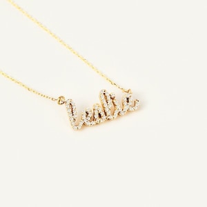 May include: A gold-toned necklace featuring a cursive nameplate pendant that spells the name Leslie. The pendant is encrusted with numerous small, clear, sparkling stones set along the entire surface of the letters, attached to a thin gold chain.