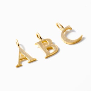 Könnte beinhalten: Drei goldfarbene Metallanhänger in Form der Großbuchstaben A, B und C. Jeder Buchstabe hat eine polierte, reflektierende Oberfläche und verfügt über einen kleinen runden Biegering an der Oberseite zur Befestigung an einer Halskette oder einem Armband.