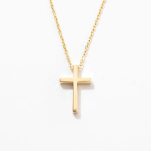 Könnte beinhalten: Eine goldfarbene Halskette mit einem schlichten, polierten Kreuzanhänger an einer zarten, passenden Kette. Das Kreuz hat klare, gerade Kanten und eine glatte, reflektierende Oberfläche, was ein klassisches und schlichtes Schmuckstück ergibt.