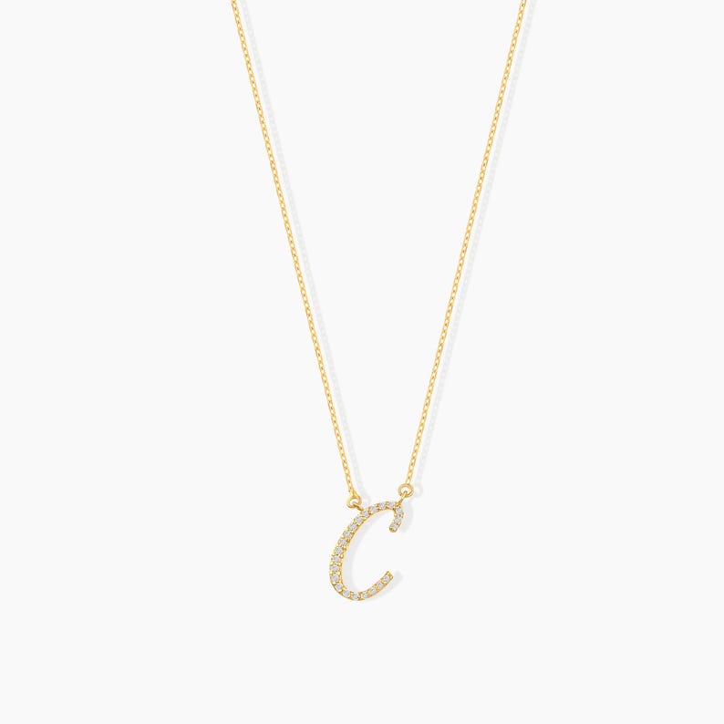 May include: A delicate gold-colored necklace featuring the letter "C" encrusted with small, sparkling gemstones. The pendant hangs from a fine chain, creating a simple yet elegant design. The necklace is displayed against a plain white background.