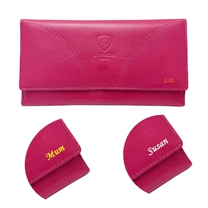 Op de afbeelding: Een magenta leren portemonnee met een klepsluiting. De portemonnee heeft decoratieve stiksels en de tekst "J. WILSON LONDON" en een logo. De naam "Liz" staat op de voorkant. Twee kleinere portemonnees worden getoond met de namen "Mum" en "Susan".