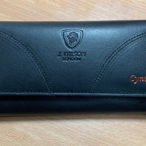 Puede incluir: Cartera de cuero negro con cierre de solapa. La cartera presenta el texto "J. Wilson London" y un escudo de león. El nombre "Cynthia" está impreso en letras color cobre en la parte delantera.