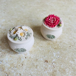 Puede incluir: Dos pequeñas cajas de cerámica blanca con diseños florales. Una caja tiene una flor rosa y la otra tiene flores blancas.