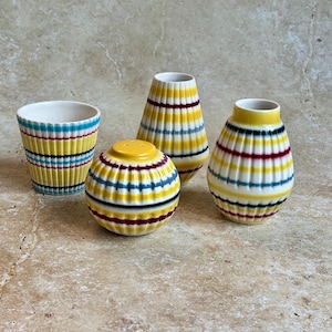 May include: Four ceramic decorative pieces with yellow, blue, black, and burgundy striped patterns. The set includes a small cup, a salt shaker, and two vases of different shapes. The pieces have a glossy finish.