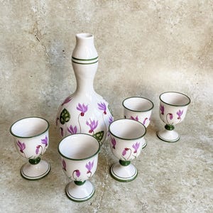 May include: A set of six white ceramic cups with green and purple floral designs. The cups have a stemmed base and are arranged around a larger white ceramic bottle with a similar floral design.