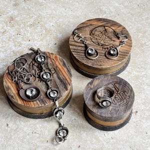 May include: A collection of silver-toned jewelry, including a necklace, bracelet, earrings, and ring, each featuring a clear, round stone. The jewelry is displayed on small, round wooden stands with engraved text. The stands have a natural wood grain finish.