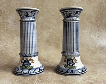 May include: Two ceramic candle holders with a column design. The holders are white with blue vertical stripes and floral accents. The base and top feature intricate blue patterns. Each holder is approximately 6 inches tall.