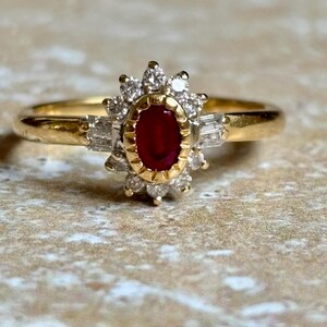 Puede incluir: Un anillo de oro con un rubí ovalado central, rodeado de pequeños diamantes y diamantes baguette. El anillo tiene un diseño clásico con una banda de oro y un halo de diamantes alrededor del rubí.
