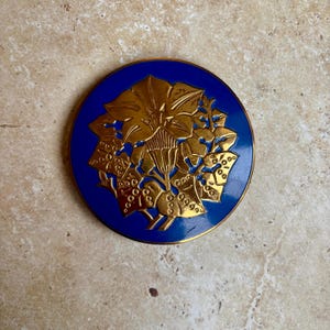 May include: A round, blue decorative object with a gold floral design. The gold design is raised and detailed, featuring flowers and leaves. The object has a gold trim around the edge and is set against a neutral background.