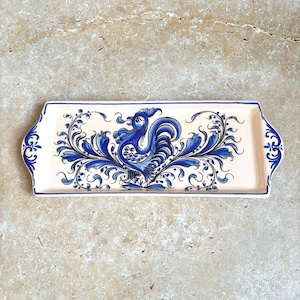 May include: A rectangular ceramic serving tray with a cream-colored base and blue hand-painted designs. The center features a stylized rooster surrounded by intricate floral scrollwork, while the side handles display matching blue floral motifs.