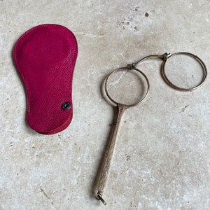 Op de afbeelding: Een antiek gouden leesbril met een scharnierend ontwerp en decoratieve details. De bril wordt geleverd met een roze leren etui.