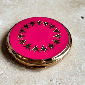 May include: A circular, gold-coloured compact with a bright pink lid. The lid is decorated with a ring of gold stars. The compact has a hinged opening and a shiny surface. A vintage cosmetic accessory.