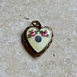 Könnte beinhalten: Ein herzförmiges Medaillon mit goldfarbenem Rahmen und cremefarbenem Innenraum. Das Medaillon zeigt zwei rosa Rosen und einen dunkelgrauen Kreis. Das Medaillon ist für ein kleines Foto oder Andenken gedacht.