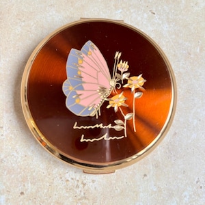 May include: A round, brown compact with a gold rim. The compact features a pink and blue butterfly with yellow accents, and yellow flowers. The compact has a gold inscription at the bottom.