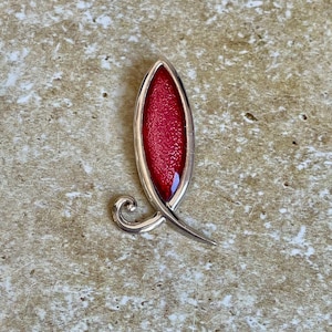 May include: A silver-toned brooch with a red, textured center. The brooch has an elongated, leaf-like shape with a swirling design element. The red center has a slightly raised, speckled appearance.
