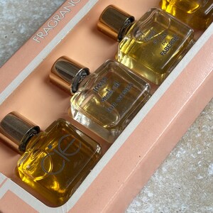 May include: A set of miniature perfume bottles in a pink box. The bottles are square with gold caps and contain a golden liquid. The text "FRAGRANCE" is visible on the box. The bottles are labeled "Jovan Musk Oil for Women" and "Cie".