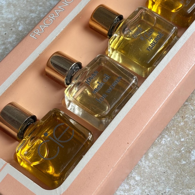 May include: A set of miniature perfume bottles in a pink box. The bottles are square with gold caps and contain a golden liquid. The text "FRAGRANCE" is visible on the box. The bottles are labeled "Jovan Musk Oil for Women" and "Cie".