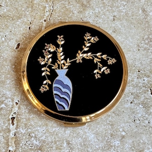May include: A round, gold-toned compact with a black enamel face. The face features a vase with a floral design in gold, red, and blue. The compact is closed and sits on a light-colored, textured surface.